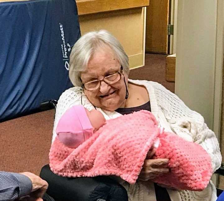 alzheimer patient sitting with glasses on smiling down at baby doll wrapped in pink blanket in her lap