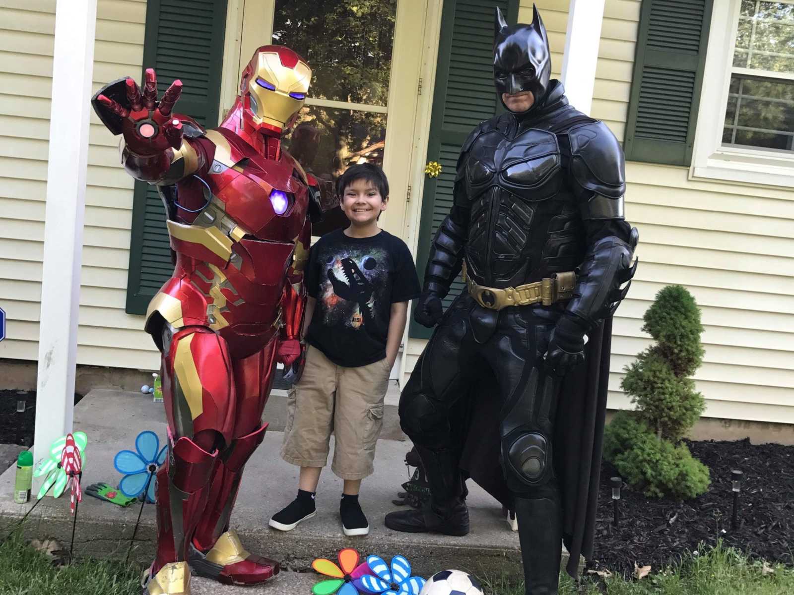 Little boy with cancer smiles on front porch with iron man on one side of him and batman on the other