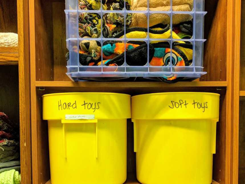 shelves with a clear container of blankets and bellow a yellow bucket that says, hard toys" and one that says, "soft toys"