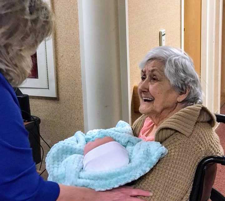 alzheimer patient in wheelchair smiles up at woman standing over her while holding baby doll wrapped in blue blanket