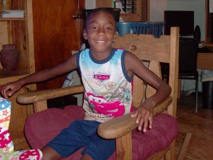 young boy sitting in wooden chair with red seat cushion wearing a disney cars tank top