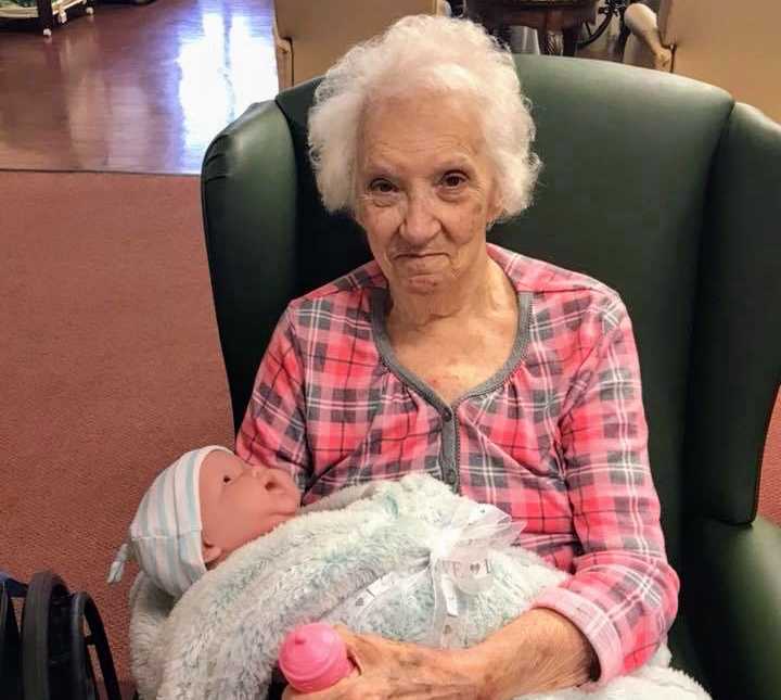 elderly woman sitting in green chair holding baby doll in arms with pink baby bottle in hand