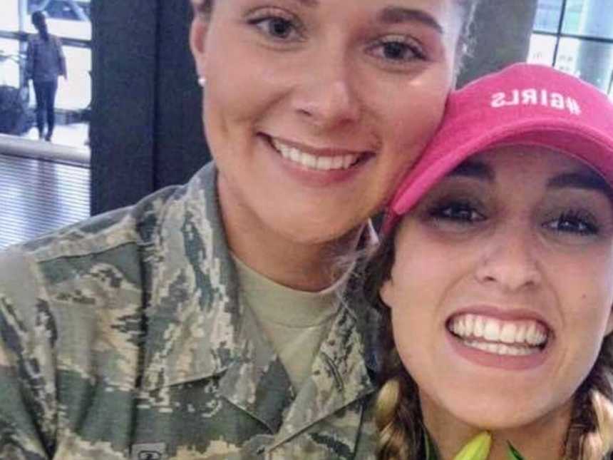 woman in army uniform smiles and rests head on another woman who is wearing a pink hat that says, "#girls" in selfie