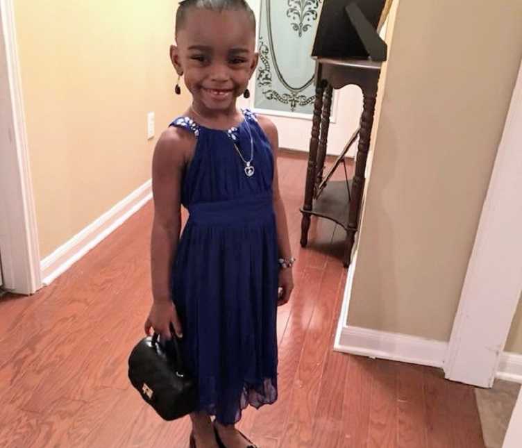 little girl smiles in hallway while wearing a purple dress and holds black purse