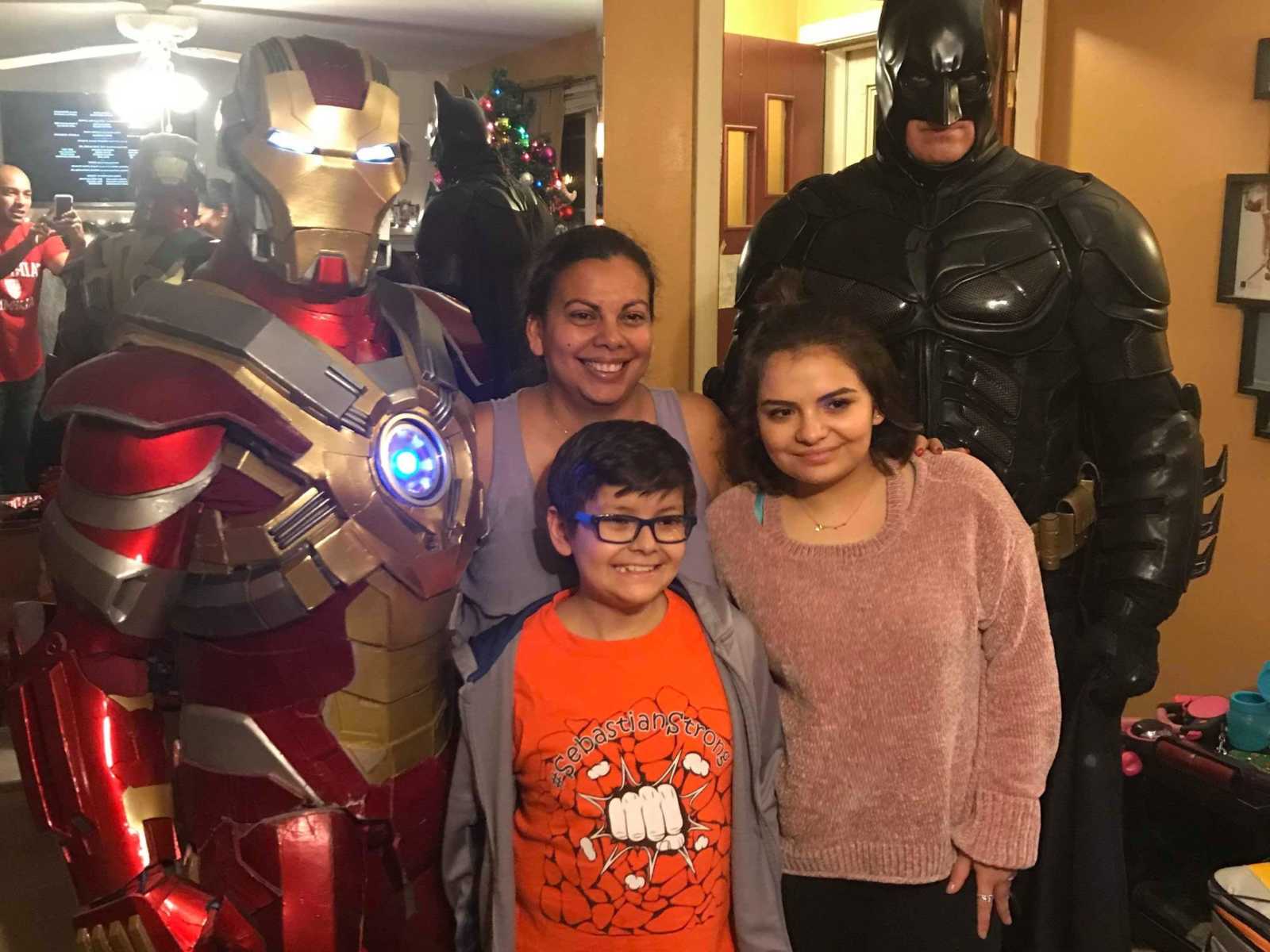 Boy with cancer smiles next to ironman, two female family members, and batman in his house