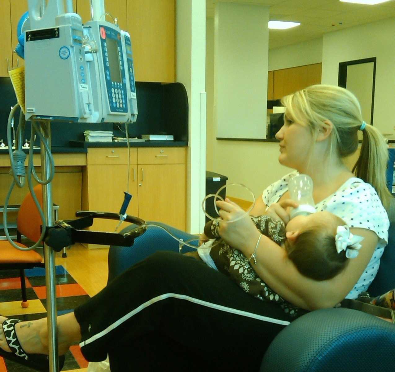 Mother holds infant with rare disease in hospital chair feeding her a bottle