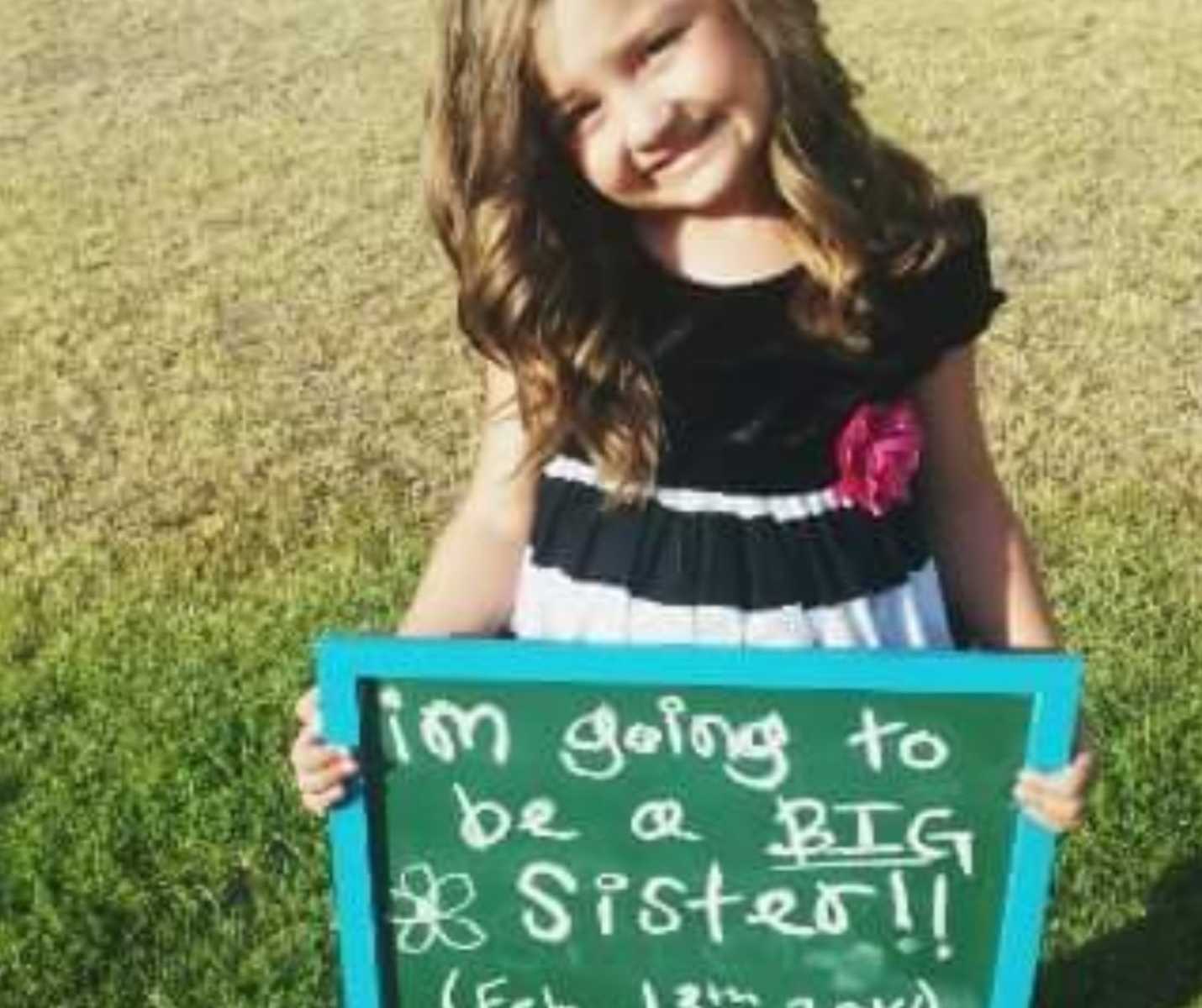 little girl in fancy dress holds chalk board that says, "i'm going to be a big sister"