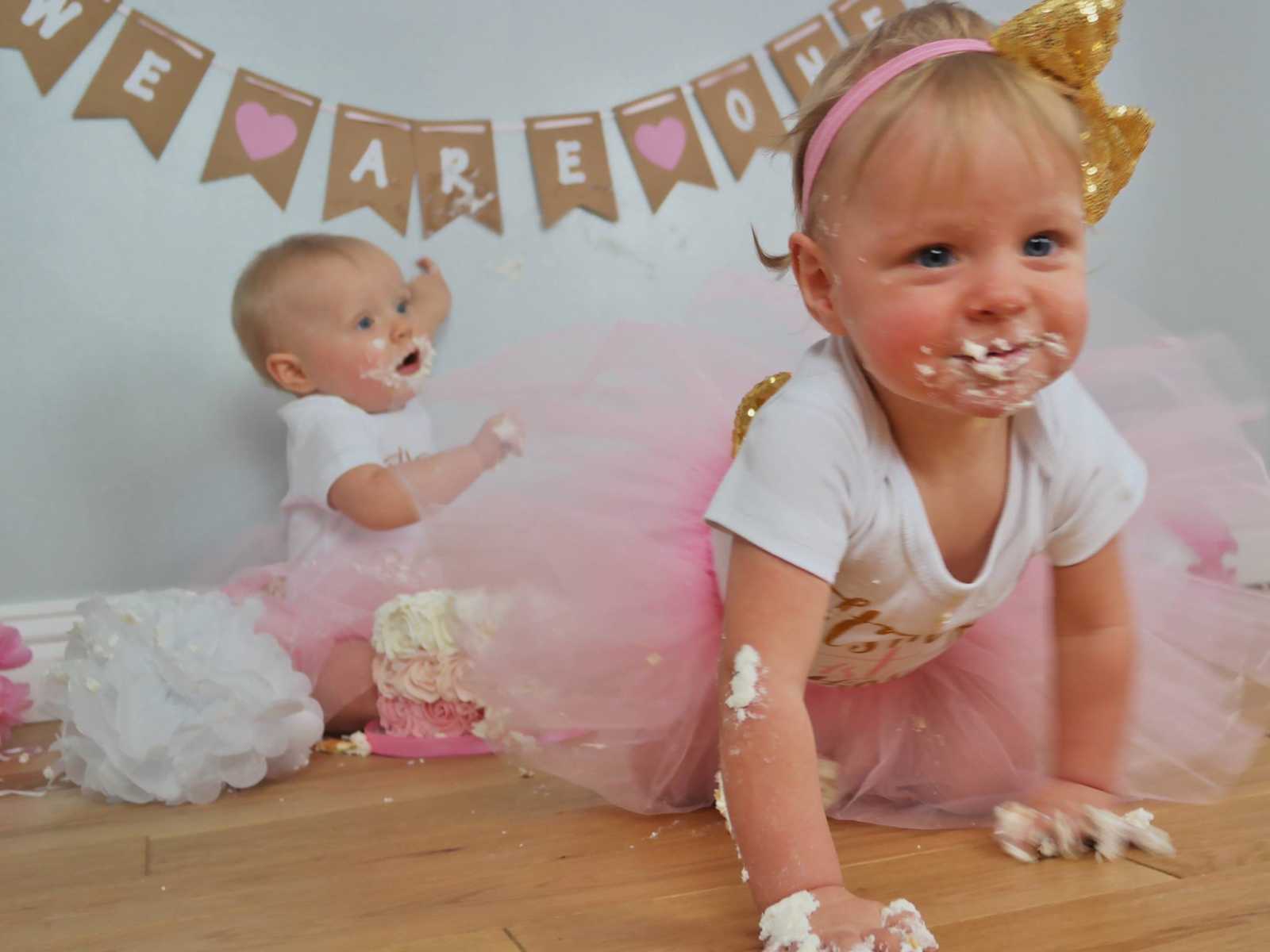 baby with birthday cake all over her crawls on table while baby in background points to banner saying, "we are one"