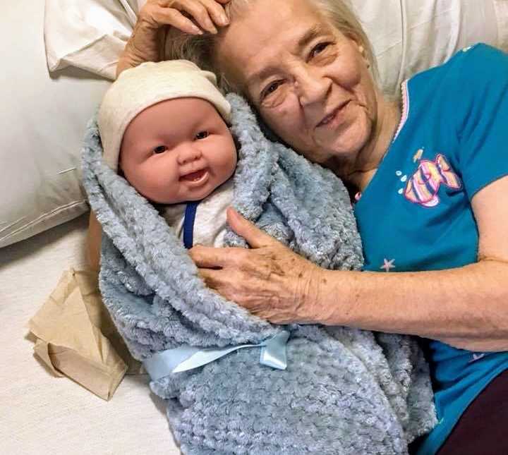 elderly woman lying in bed smiling with her hand on baby dolls chest lying next to her