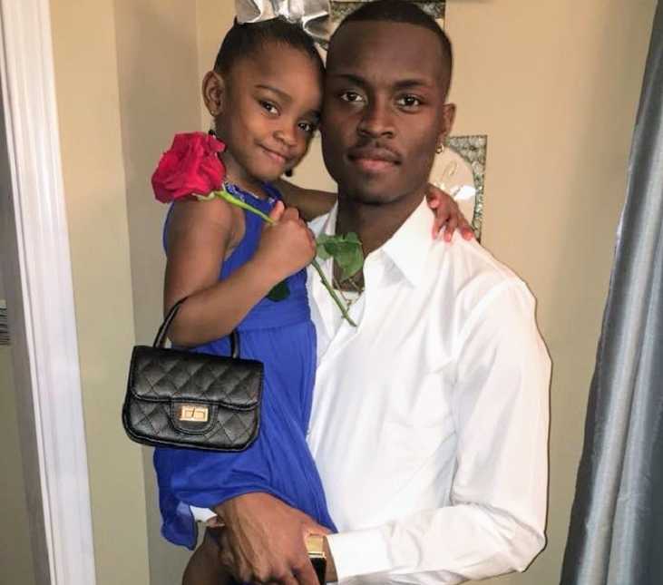 father smiles while holding daughter in purple dress with black purse and red rose in hand