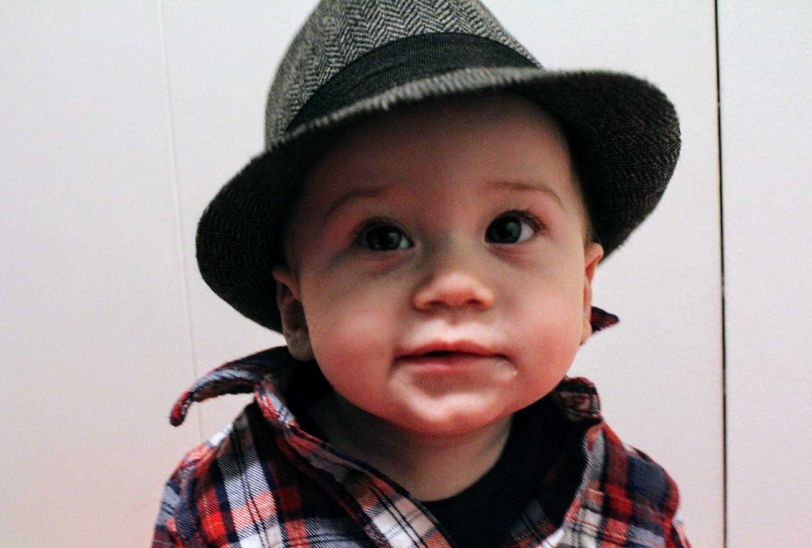 close up of infants face who is wearing a black and white herringbone fedora and red white and blue flannel