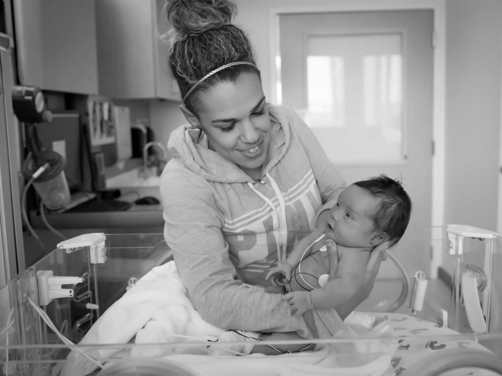 mother smiles while leaning over newborn hospital bed to pick up daughter with down syndrome