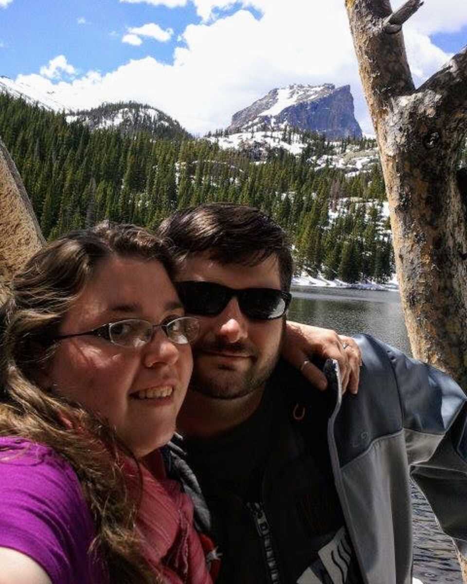 Husband and wife take selfie in front of body of water, trees, and mountains