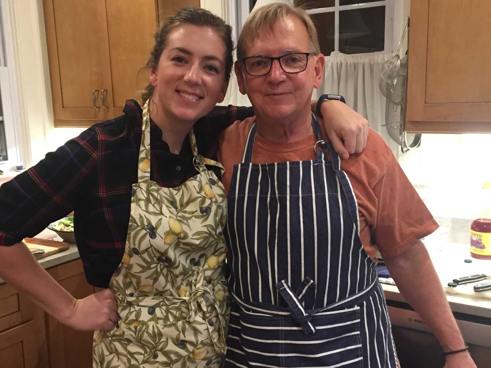 daughter stands in apron with on hand on hip and the other around dads shoulder who is also wearing apron