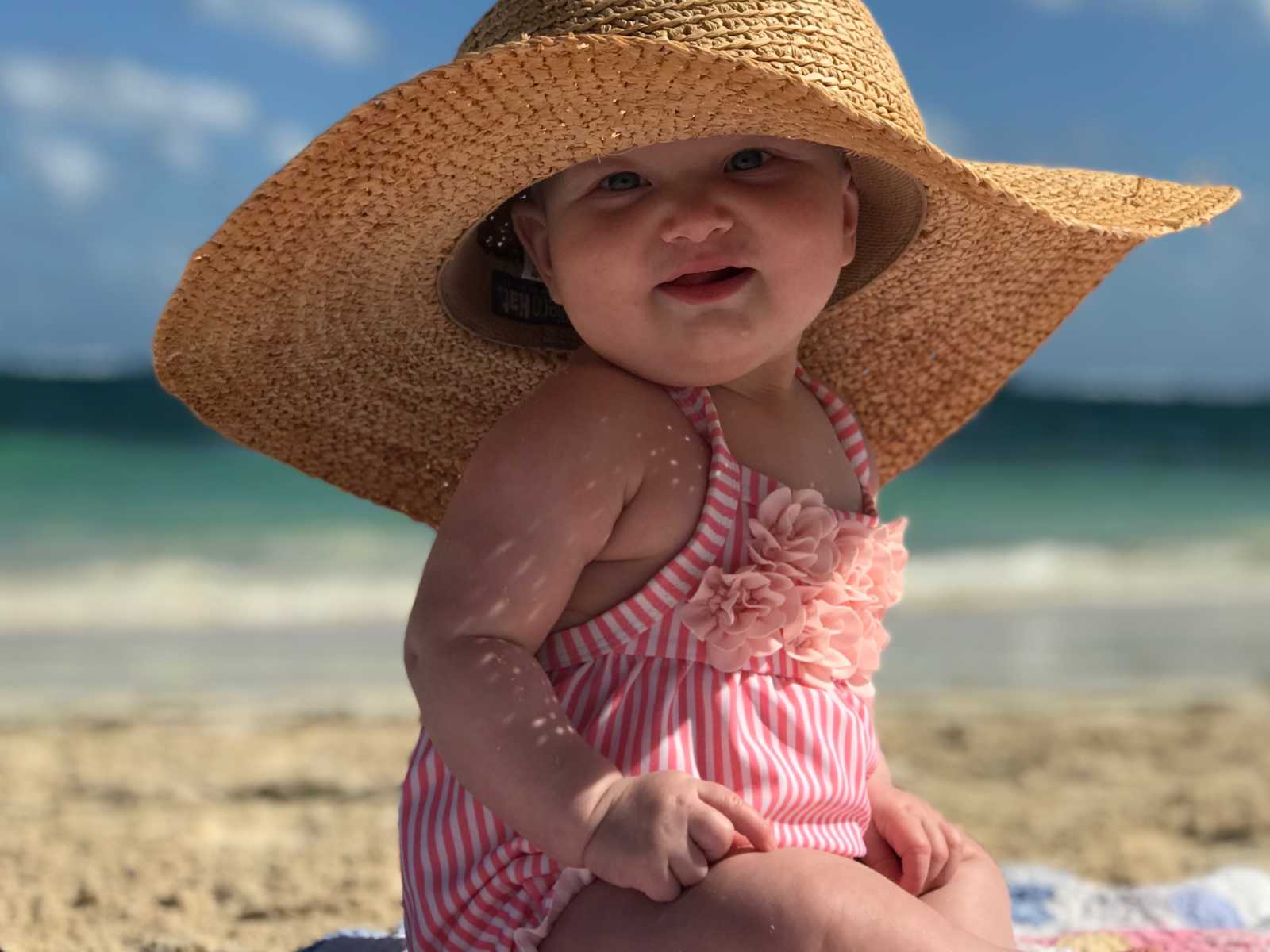Baby whose mother has postpartum thyroid condition sits on beach with mothers beach hat on smiling