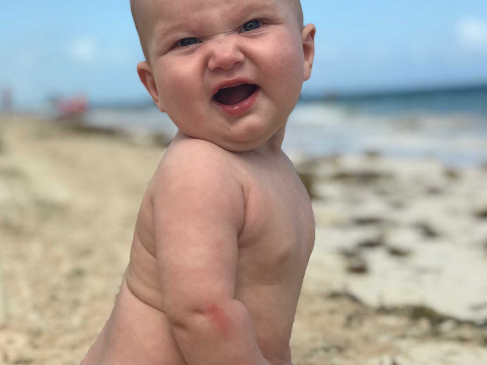 Baby whose mother has postpartum thyroid condition without bathing suit on makes funny face at beach