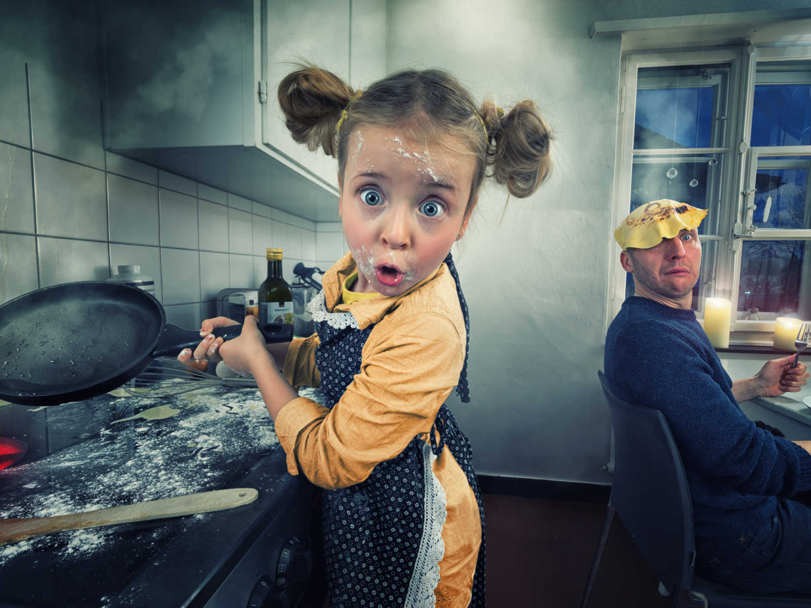 Young girl stands in animated messy kitchen with frying pan in hand while dad has pancake on his head in background