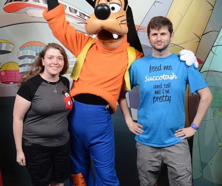 Husband and wife who lost combined amount of 160 pounds stand on either side of Goofey at Disneyworld