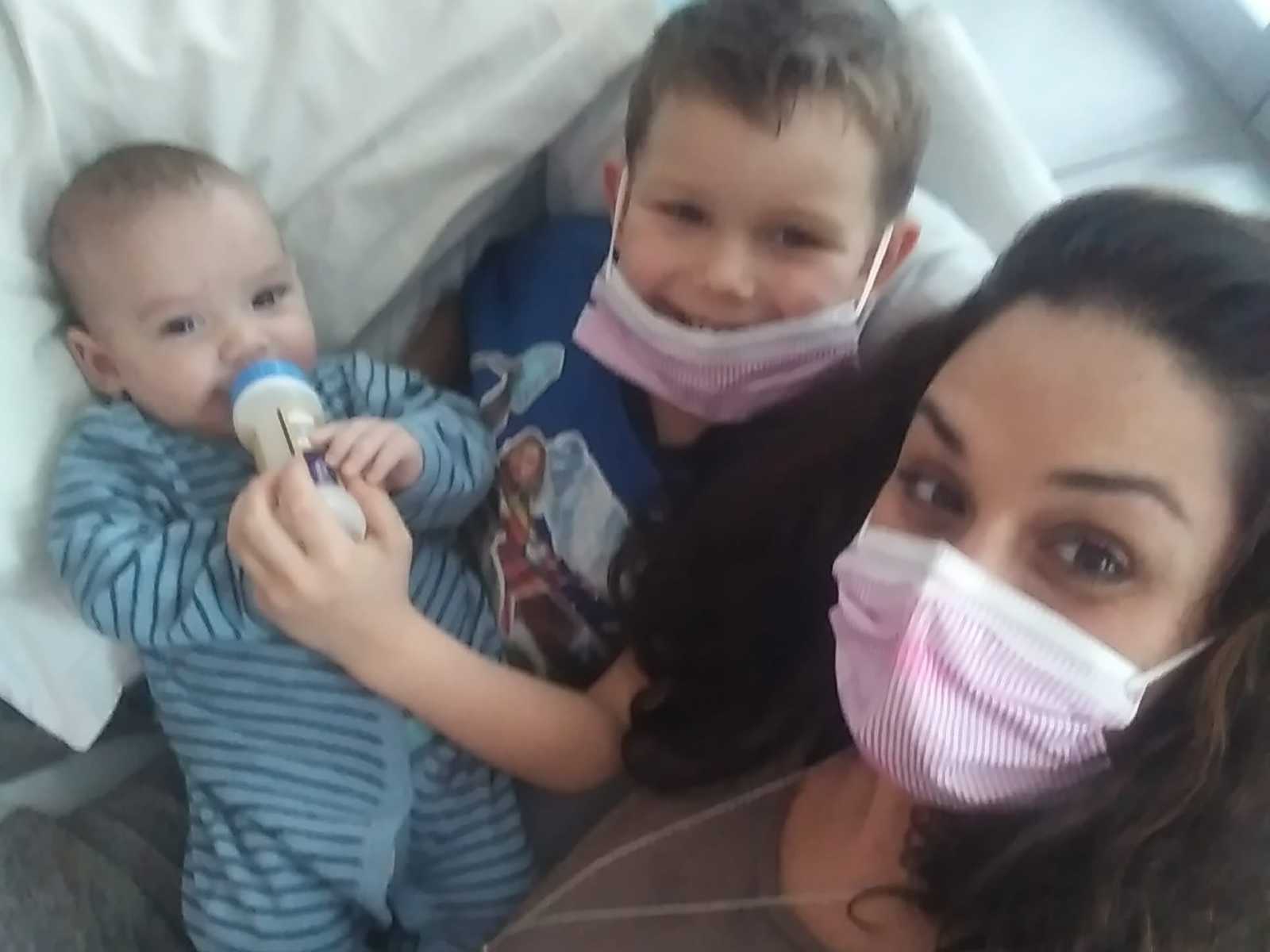 Mother wearing a mask takes selfie of son who donated bone marrow to baby siblings feeding little brother