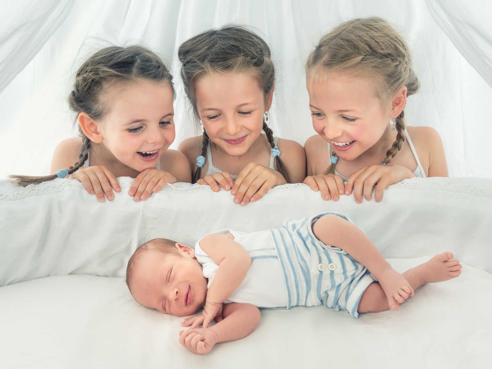 three girls smiling over their newborn sibling who is sleeping in white crib