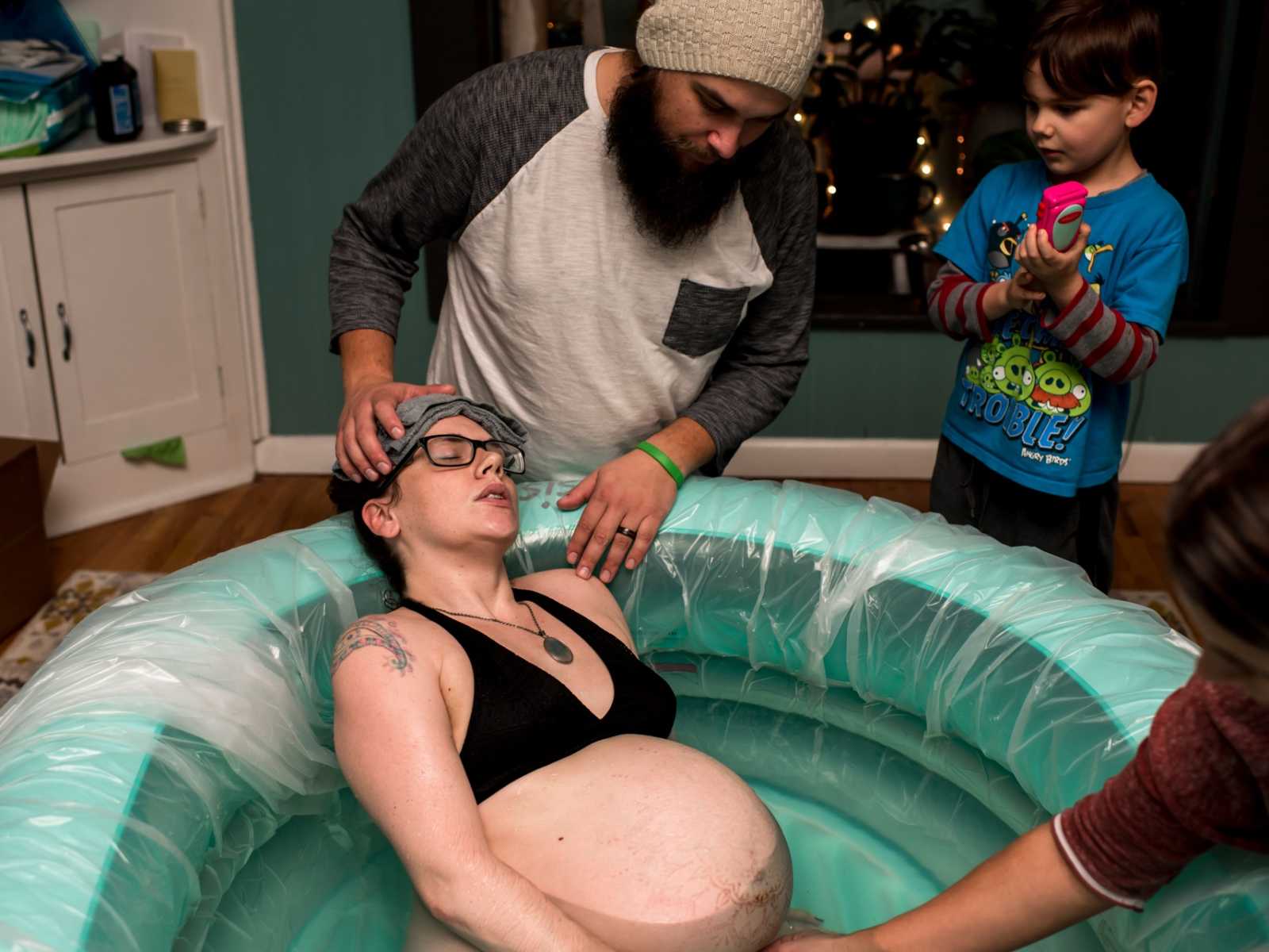 Pregnant woman leaning back in birthing pool while husband holds towel on forehead and toddler son watches