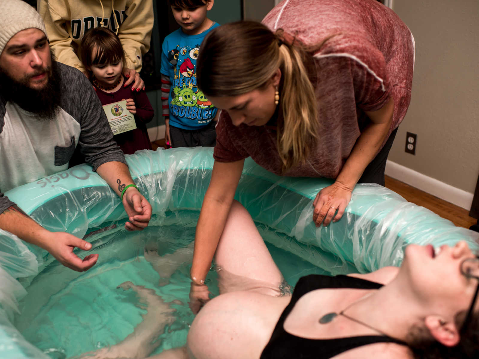 Midwife reaching into birthing pool to help woman give birth while family watches