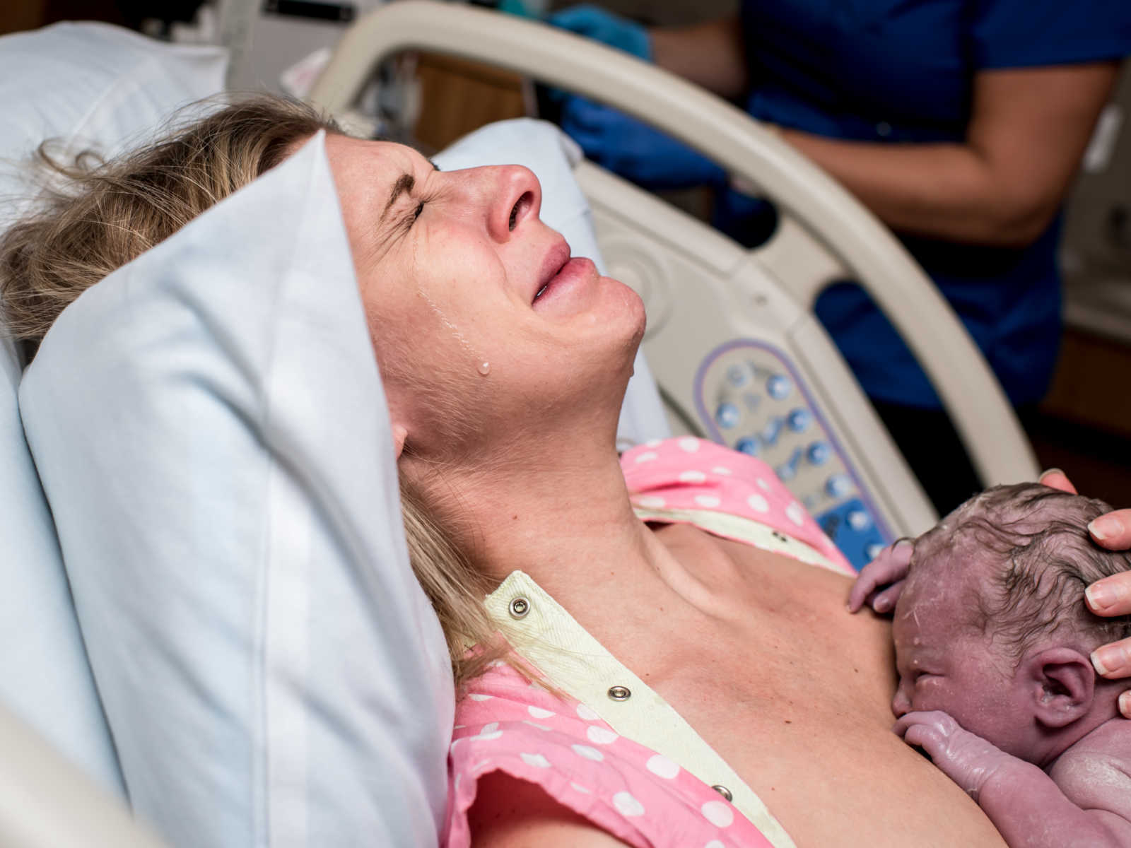 women holds newborn baby to chest as she leans head back on pillow with tears down her face