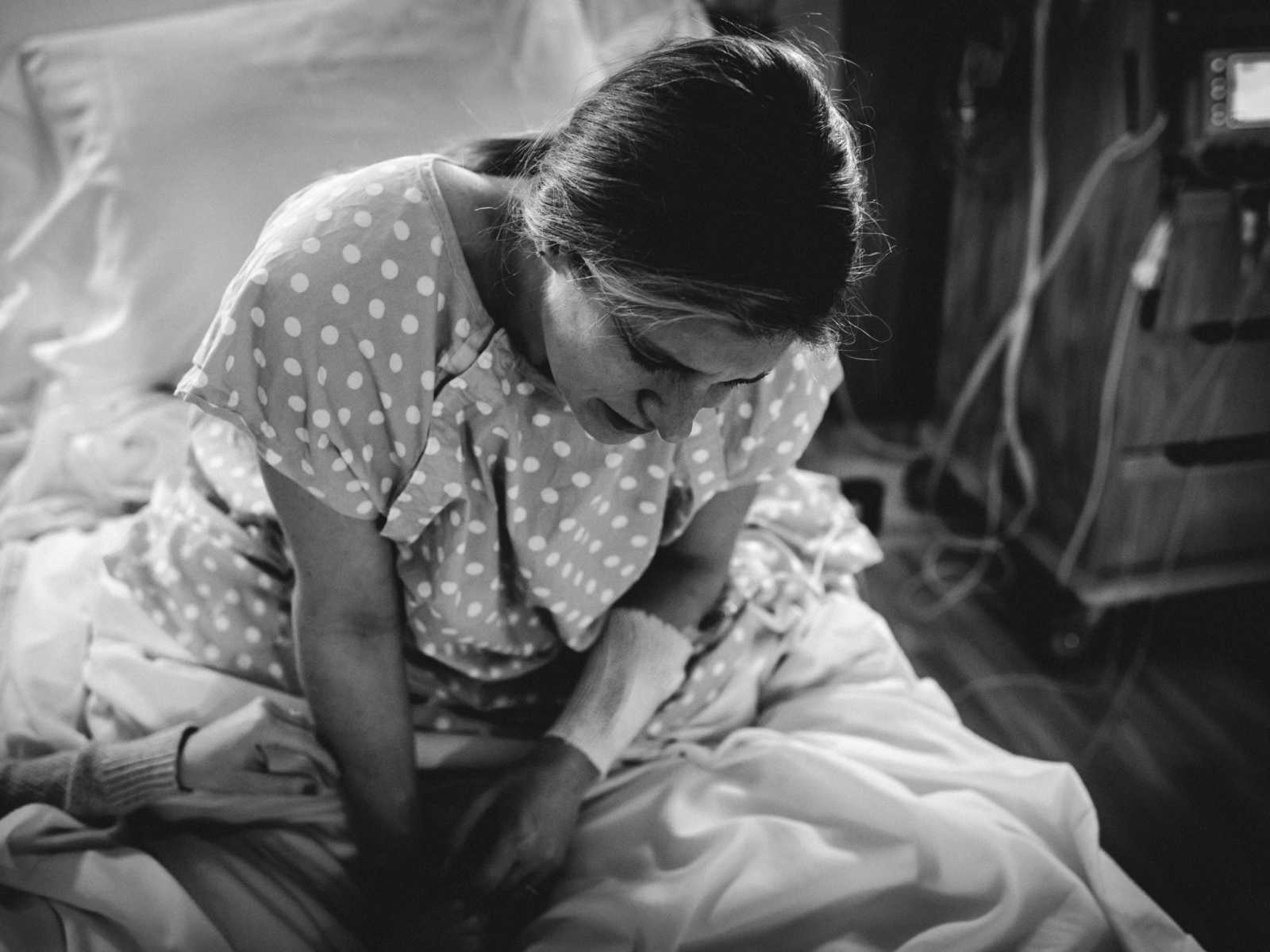 pregnant woman in polka dot hospital gown sits up in hospital bed in agony while a hand reaches out to her