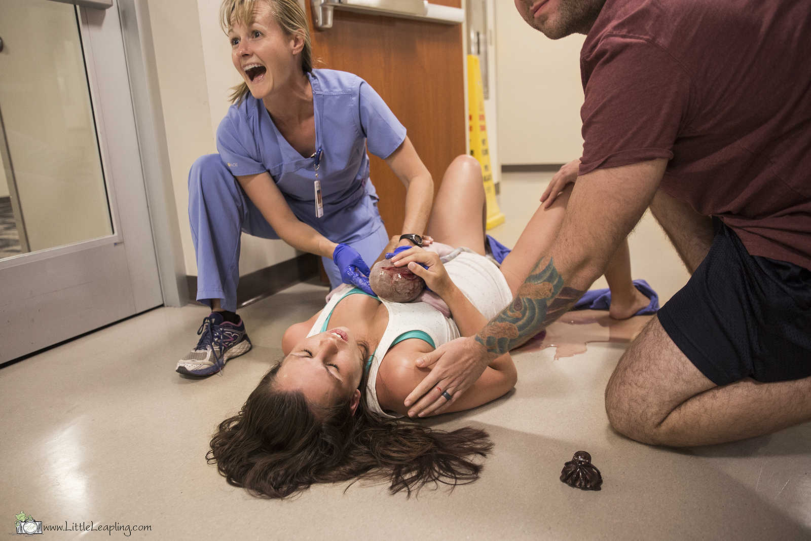 Nurse and husband kneel next to woman who holds new born baby on hospital floor in a pool of blood