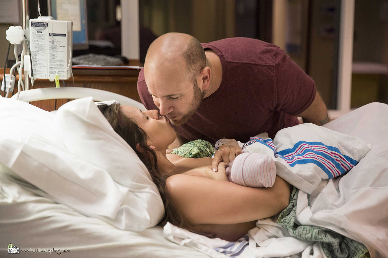 husband leans to kiss wife who is breastfeeding their newborn baby in hospital bed