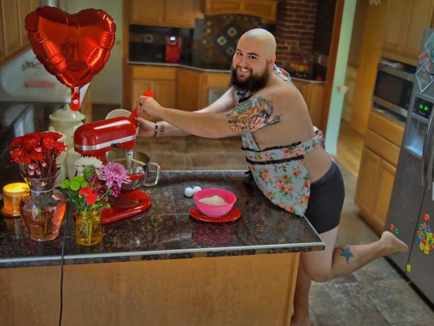 shirtless husband wearing apron and boxers smiles while pouring ingredients into kitchen aid mixer next to flowers 