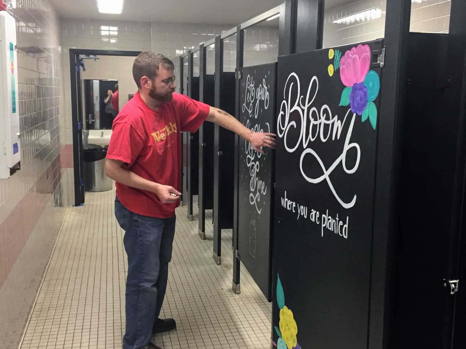 man standing in public restroom holding stall door containing inspirational message saying, "bloom where you are planted"