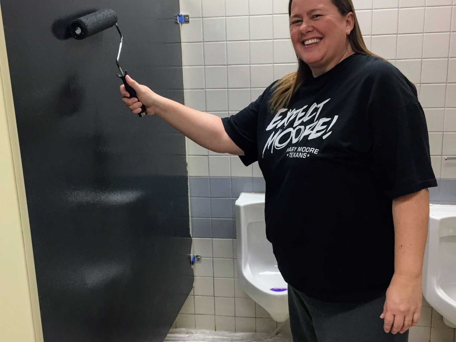 woman smiling in public restroom in front of urinals as she paints stall door black