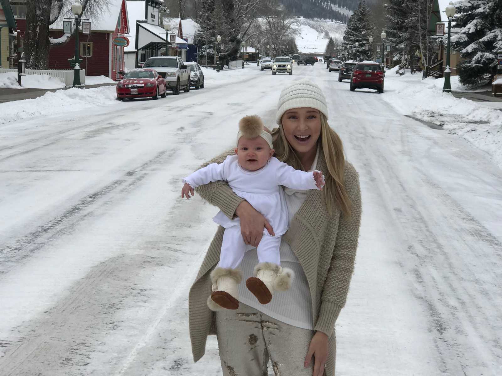 Woman with postpartum thyroid condition holds baby in middle of snowy road smiling