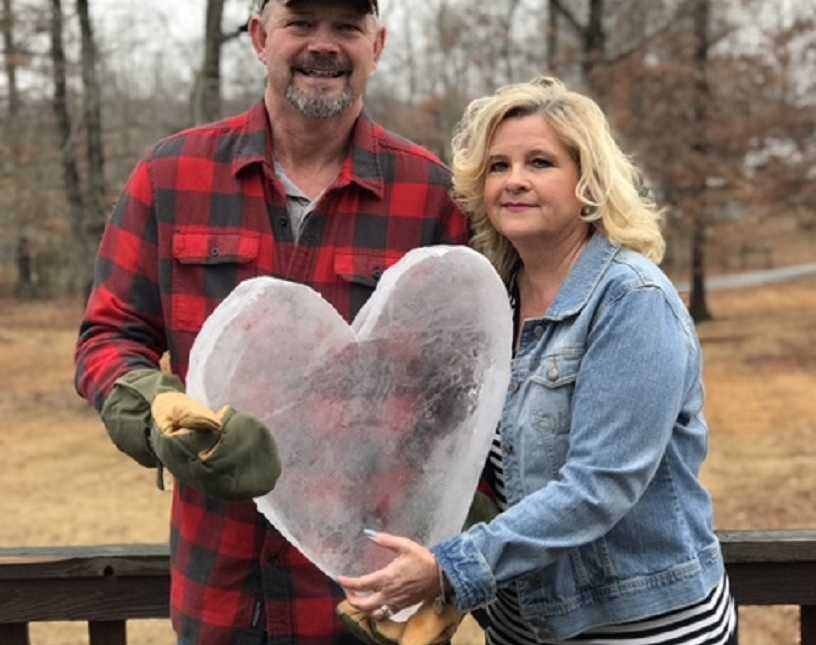 husband stands next to wife smiling as they hold piece of ice shaped as a heart