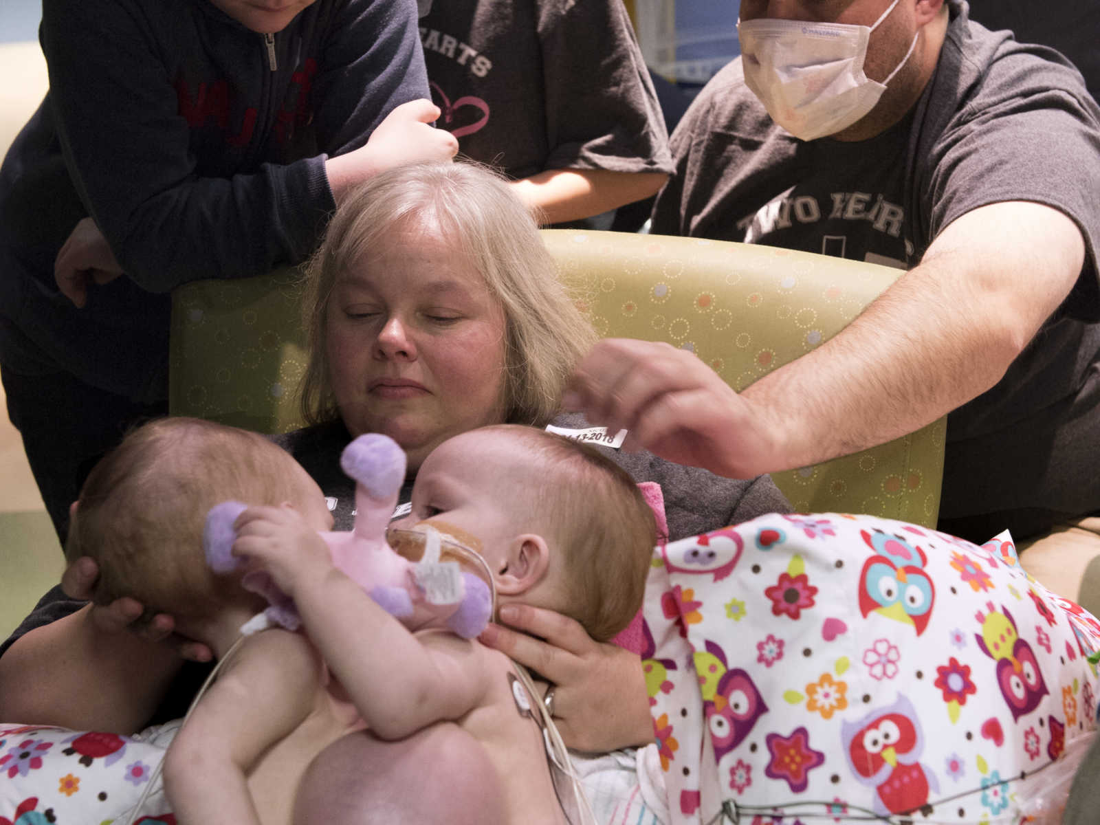 mother holding conjoined twins in lap while man with mask reaches over her shoulder to touch twins next to two others