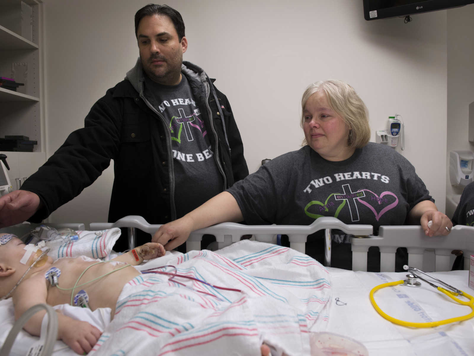 mom and dad standing at side of hospital bed reach arms out to touch baby after surgery 