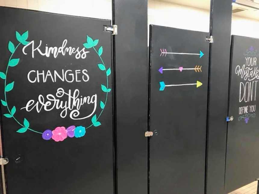 black bathroom stall with, "kindness changes everything" surrounded a vine and flowers next to door with three arrows on it