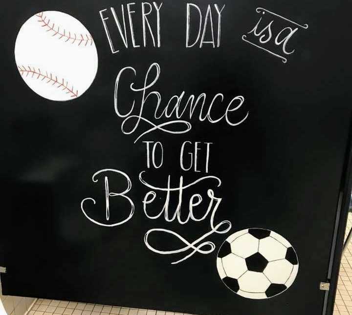 bottom of black stall bathroom door with quote, "every day is a chance to get better" on it with baseball and soccer ball