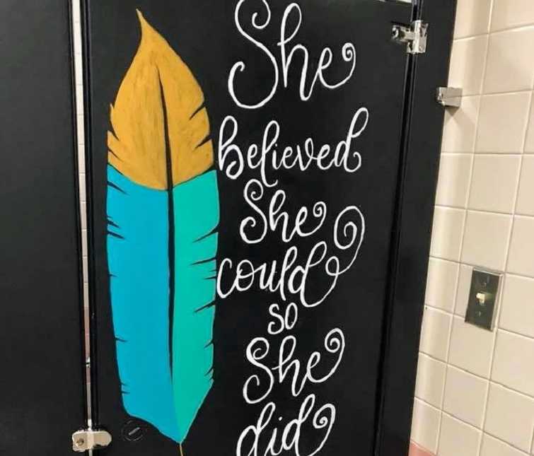 bathroom stall door with yellow blue and green feather on left and quote, "she believed she could so she did on right
