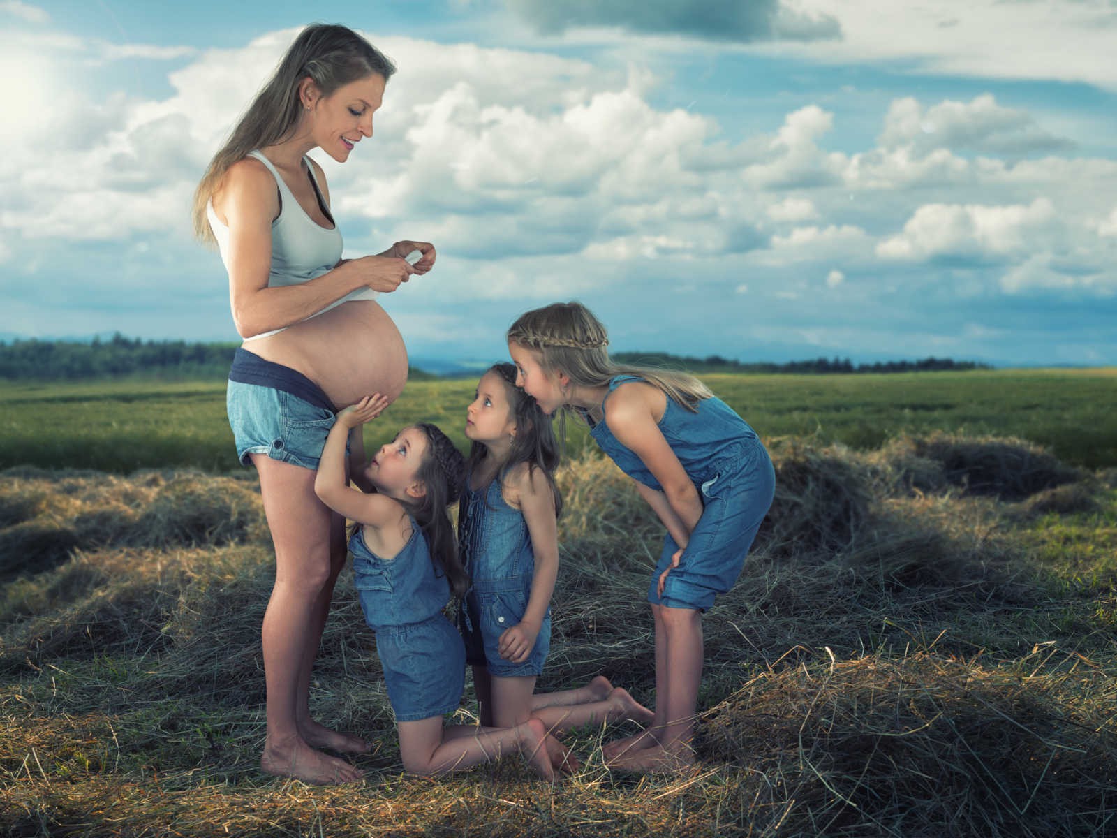 three siblings crouch down next to pregnant mothers stomach while she looks down at her daughters