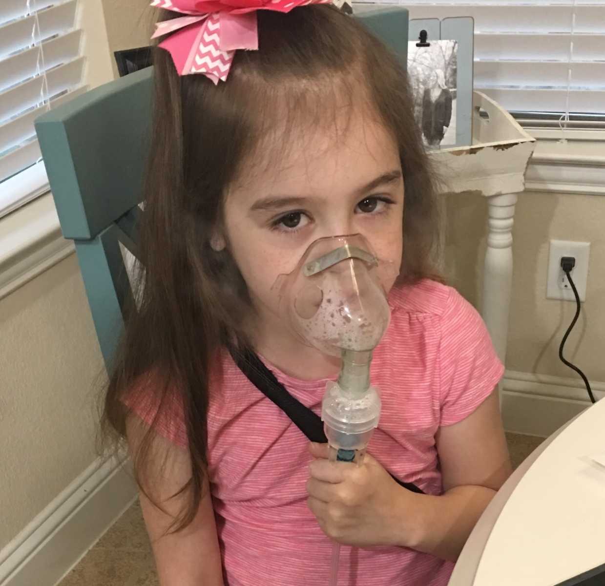 Little girl with rare disease sits at table while holding an oxygen mask to her mouth