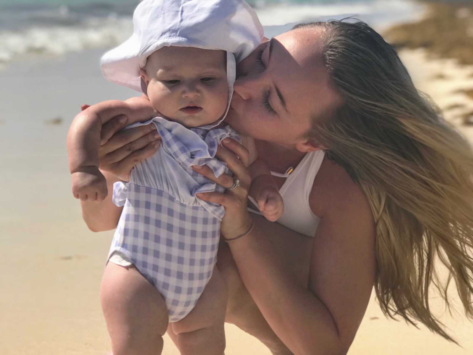 woman who struggles with postpartum thyroid condition holds up baby and kisses her cheek on beach