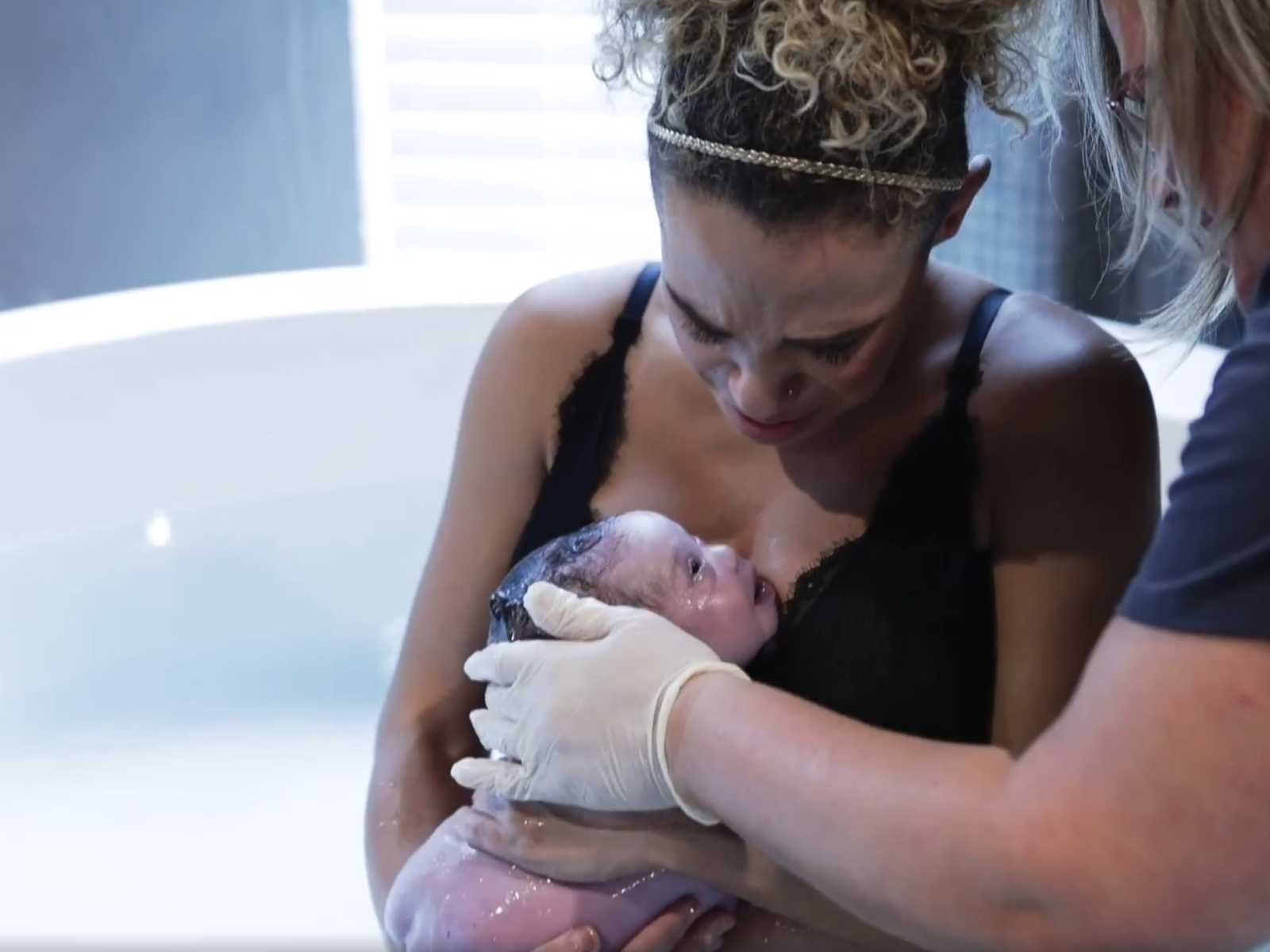 mother looking down at wet newborn next to woman who has hand in white latex glove placed on newborns head