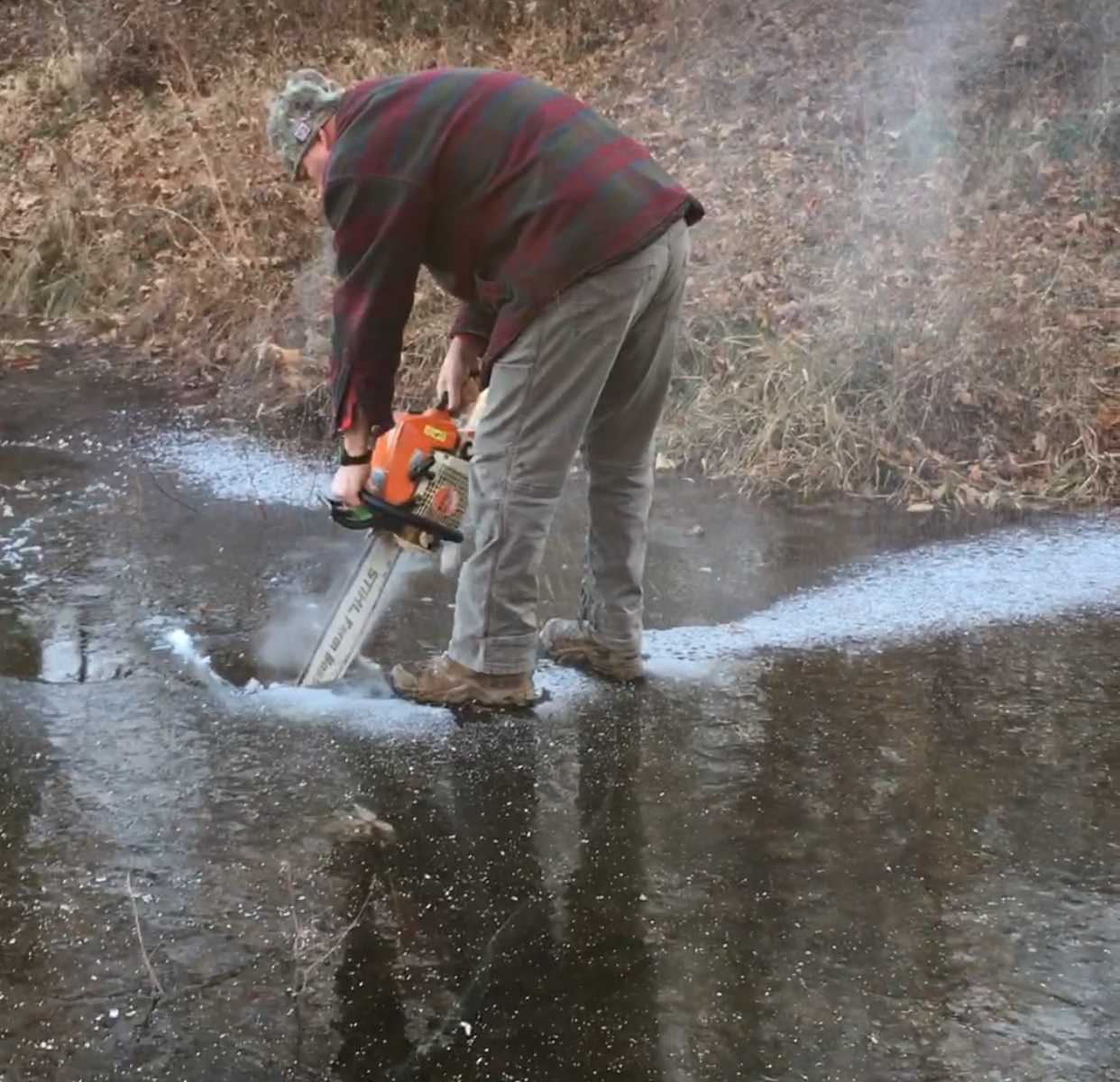 man leaning over with chain saw to cut through frozen body of water