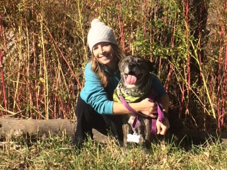 owner sits on ground with arm around adopted tongue who has its tongue out in the woods