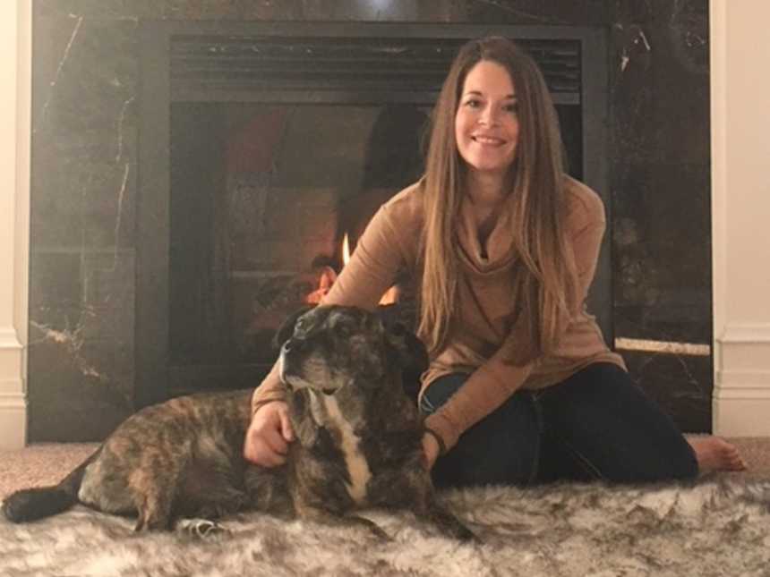 adopted dog sits in front of fire place while owner sits smiling next to it with arm placed on dogs back