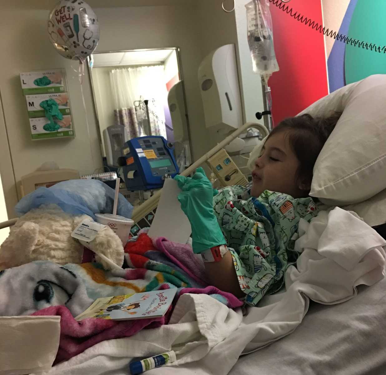 Toddler with rare disease lying in hospital bed with fuzzy blankets, books, and stuffed animals