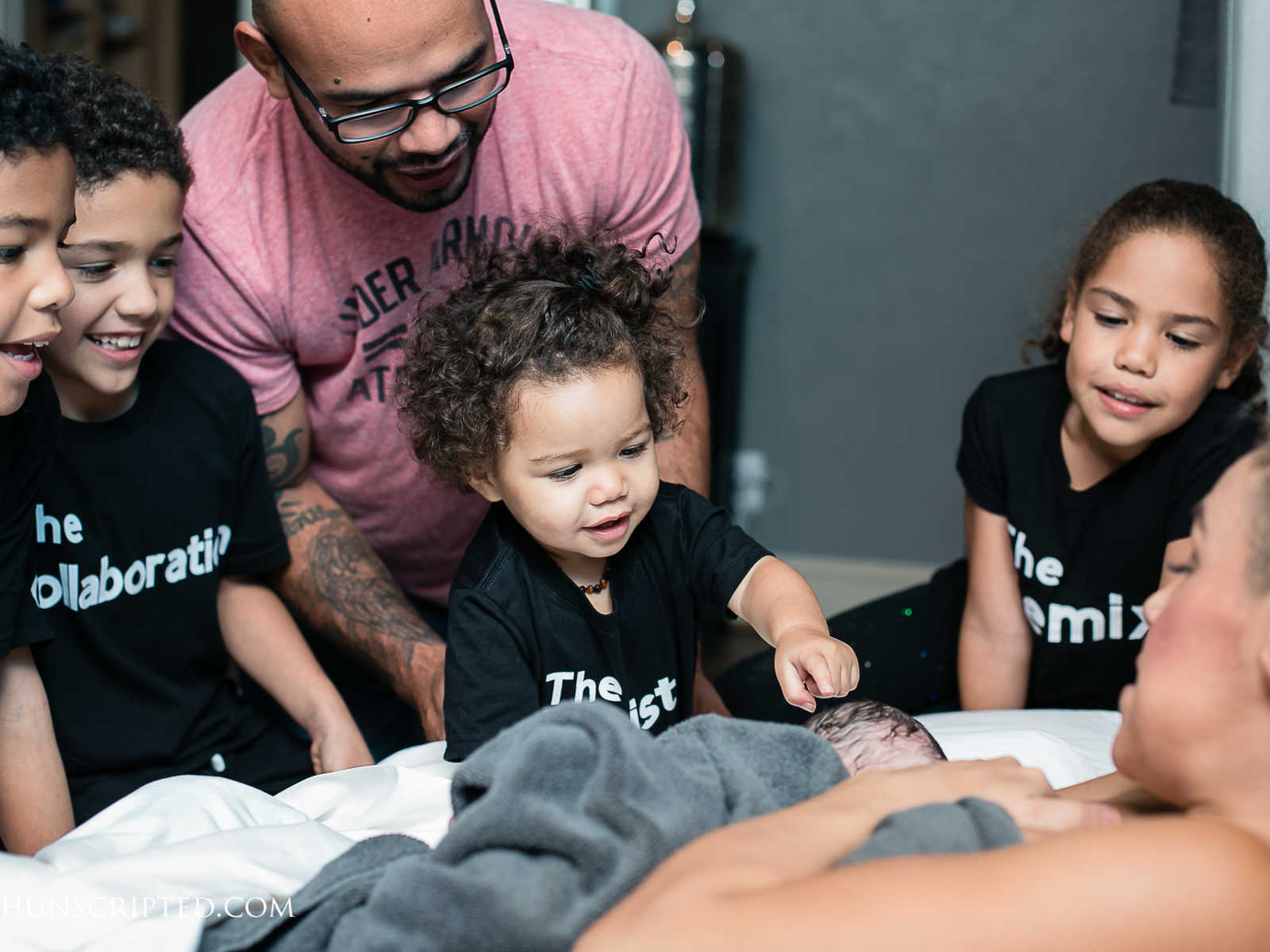 father and two sons and daughters lean over to look at newborn baby lying next to mother 