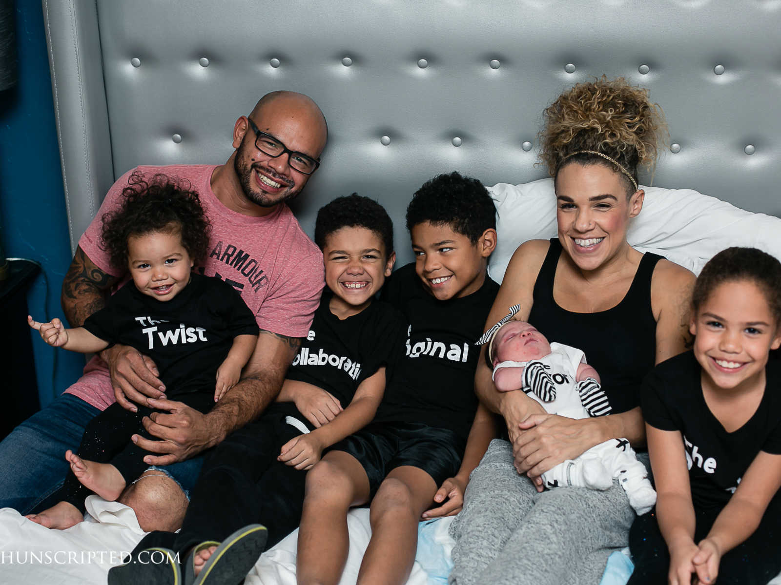 mother smiles in bed with newborn in lap next to husband and other two sons and daughters